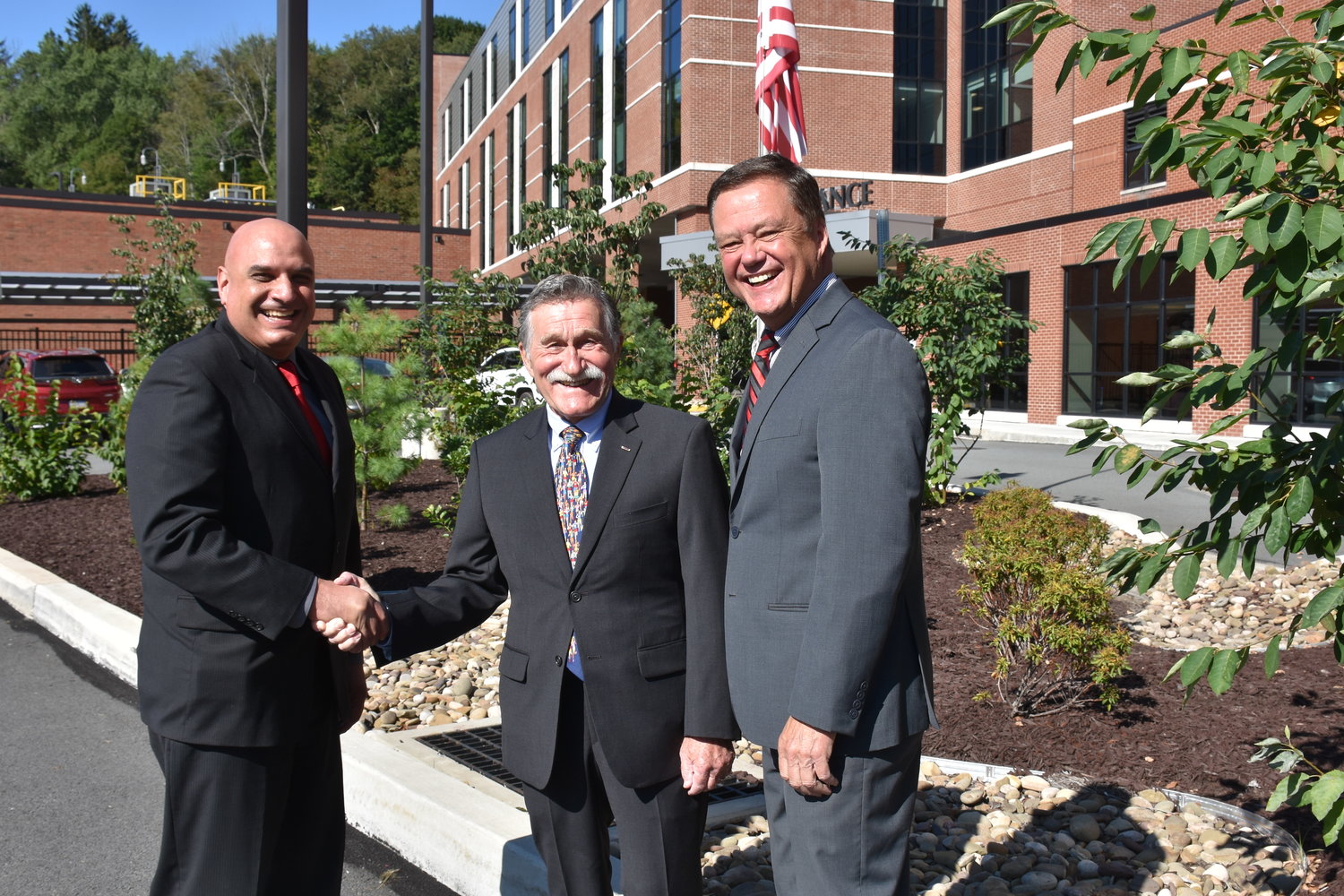 James Pettinato, left, has been chosen as the new chief executive officer at the Wayne Memorial Health System. He is congratulated by Board Chairperson Hugh Rechner, center. Pettinato will succeed David L. Hoff, right. The flag behind them is at half-staff in honor of the 13 service members and others killed recently in Afghanistan.